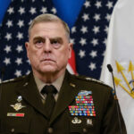 A military leader in a decorated uniform standing in front of U.S. flags