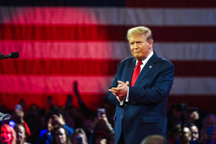 A man in a suit stands in front of an American flag, applauding at a rally