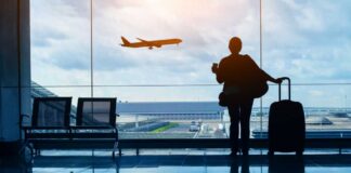 Silhouette of a traveler holding a coffee cup and standing next to a suitcase, looking out at an airplane taking off from the airport