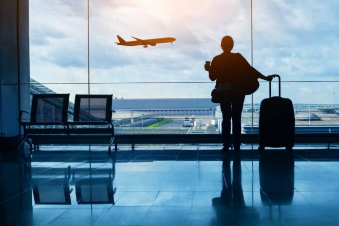 Silhouette of a traveler holding a coffee cup and standing next to a suitcase, looking out at an airplane taking off from the airport
