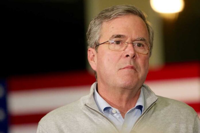 Man in a sweater speaking at a political event with an American flag backdrop