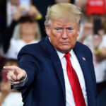 A man in a blue suit and red tie gestures while speaking at a rally