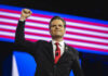A man in a suit raises his fist in celebration on stage with an American flag backdrop