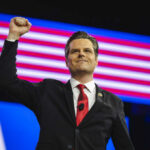 A man in a suit raises his fist in celebration on stage with an American flag backdrop