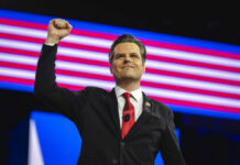 A man in a suit raises his fist in celebration on stage with an American flag backdrop