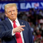 A man in a suit gestures while speaking at a political rally with a cheering crowd in the background