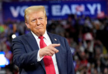 A man in a suit gestures while speaking at a political rally with a cheering crowd in the background