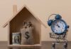 A wooden house model with money and a clock in a shopping cart