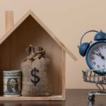 Tariffs Slam Homeowners — Insurance Costs Soar! A wooden house model with money and a clock in a shopping cart