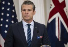 A man in a suit speaking at a podium with flags in the background