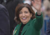 A smiling woman in a green coat waving at an outdoor event