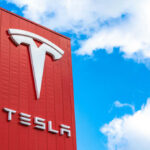 Tesla logo on a red corporate sign against a blue sky with clouds
