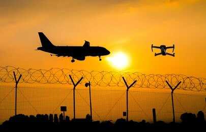 An airplane and a drone flying against a sunset backdrop with a barbed wire fence
