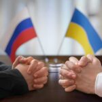 Clasped hands of two individuals with Russian and Ukrainian flags in the background