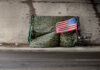 Outrage as NYC Bets $4.2B in Budget Turmoil A makeshift shelter draped with an American flag next to a shopping cart on a city street