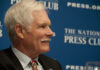 A man with gray hair and a mustache smiling during a press conference