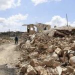 Person walking past rubble and destroyed buildings on a dirt road