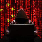 A hooded figure sitting in front of a laptop with a red digital background