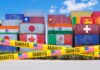 Stacked shipping containers with national flags behind yellow tariffs tape
