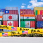 Stacked shipping containers with national flags behind yellow tariffs tape