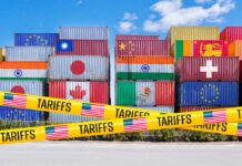 Stacked shipping containers with national flags behind yellow tariffs tape
