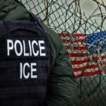A police officer in a tactical vest stands near a barbed wire fence with an American flag in the background