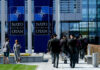 NATO Commits To Massive Defense Spending Military personnel walking past NATO banners with two women conversing in the foreground