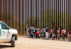 DOJ Exposes Sick Border Smuggling Plot Group of families and children walking near a border wall with a customs vehicle nearby