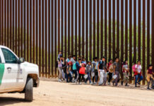 DOJ Exposes Sick Border Smuggling Plot Group of families and children walking near a border wall with a customs vehicle nearby