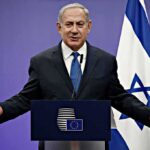 man in suit speaking at a podium with Israeli flag