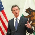 FBI Mole Penetrates Newsom’s Inner Circle A government official delivering a speech with flags in the background