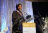 Minnesota Raid Tangles Omar’s Family Woman in hijab speaking at a podium on a decorated stage
