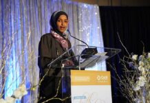 Minnesota Raid Tangles Omar’s Family Woman in hijab speaking at a podium on a decorated stage
