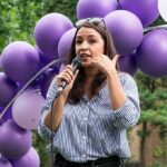 AOC’s Bold Move Rattles 2028 Field Woman speaking into a microphone at an outdoor event with purple balloons