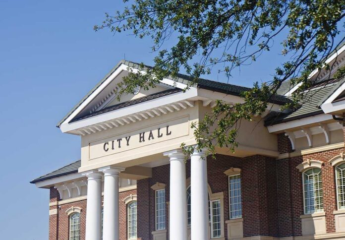 A,Classic,Brick,Town,City,Hall,Through,The,Trees Exterior view of a city hall building with columns and a clear blue sky