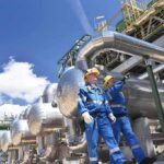 Two industrial workers walking under large metal pipes