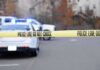 Police tape cordons off a street with patrol cars and flashing lights in the background