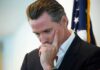 FBI Raids Expose Homeless Cash Heist Gavin Newsom in a pensive pose with hand on face near a U.S. flag