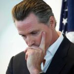 Gavin Newsom in a pensive pose with hand on face near a U.S. flag