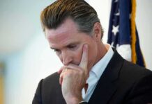 FBI Raids Expose Homeless Cash Heist Gavin Newsom in a pensive pose with hand on face near a U.S. flag