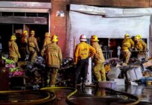 Citywide Inferno Tied To Encampments Firefighters work outside a damaged warehouse with debris and hoses