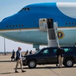 Security detail near blue and white aircraft with staircase and SUV