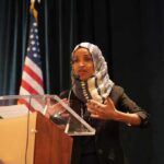 Immigration Fraud Bomb Ticks Under Omar Ilhan Omar speaking at a podium with a microphone
