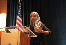 Immigration Fraud Bomb Ticks Under Omar Ilhan Omar speaking at a podium with a microphone