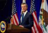 Newsom’s Blitz: Gun Owners On Notice Man in suit speaking at a podium with American and California flags