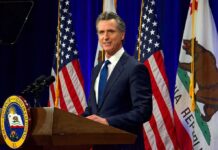 Newsom’s Blitz: Gun Owners On Notice Man in suit speaking at a podium with American and California flags